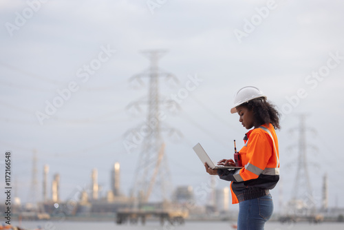 A female engineer wearing a white safety helmet and orange reflective jacket stands outdoors holding a clipboard, observing and analyzing a power plant facility with electrical towers in the backgroun