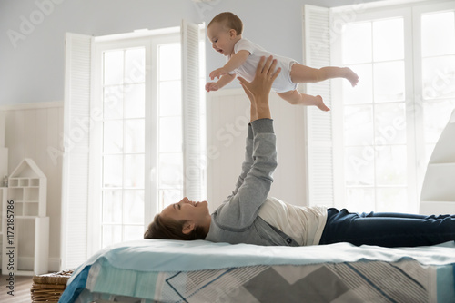 Imaginary flight. Carefree happy young mother play interact with cute lovely baby at light bedroom lying on back on bed hold laughing infant in air on stretched arms having fun at playtime. Copy space photo