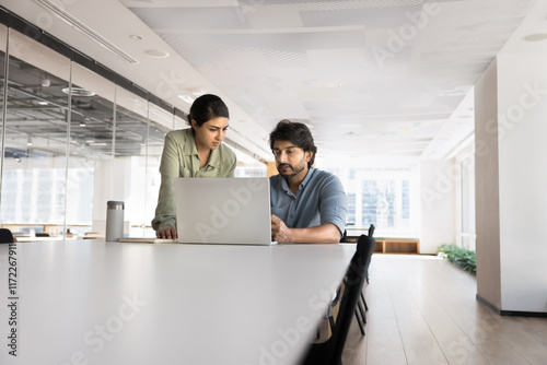 Couple of young Indian professional colleagues working on online project together, talking at laptop, meeting at large table in open office, discussing cooperation. Low angle shot with copy space