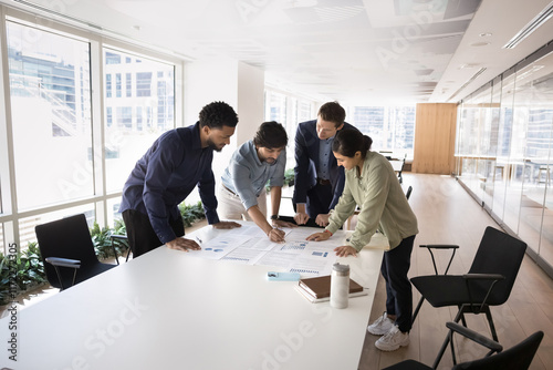 Multiethnic project managers reviewing marketing reports together, discussing statistic paper documents on large meeting table, cooperating on project strategy in open office, co-working space