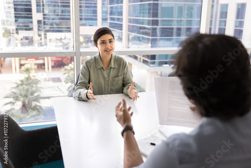 Positive beautiful Indian business professional woman meeting with male partner at table, talking to lawyer, advisor, discussing paper legal document. Job candidate speaking to employer on Interview