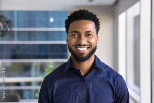 Cheerful handsome young African American businessman posing for head shot in office, looking at camera with toothy smile. Confident successful Black business leader man, startup manager portrait photo