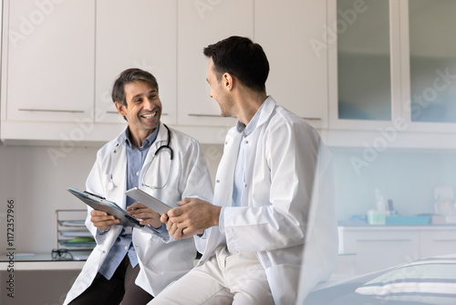 Happy successful middle aged doctor men holding tablet, medical documents, talking, discussing job, successful practitioner career, smiling, laughing. Medic workers chatting on work break
