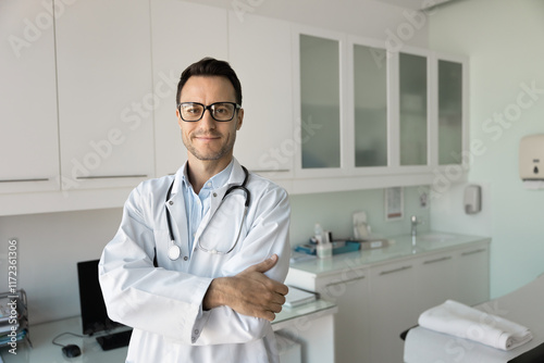 Positive confident young doctor man in glasses and white coat standing in clinic examination room with couch, looking at camera with arms folded, smiling, posing for medical professional portrait