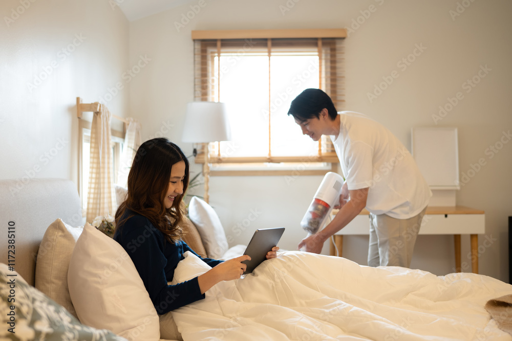 Fototapeta premium Cheerful woman using digital tablet on bed while her husband vacuuming floor beside