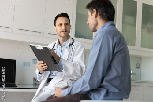 Caring young male doctor meeting with patient in clinic examination room, asking about symptoms, making records in paper documents, writing notes, talking to patient. Low angle candid shot