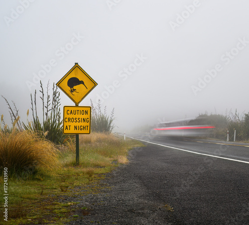 Obraz na płótnie Kiwi Crossing at Night road sign by the side of road. A car light trail speeding pasting the sign in the fog. Tongariro National Park. New Zealand. w salonie