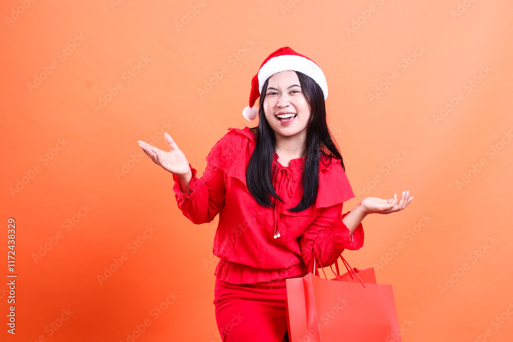 Obraz premium expression of young woman wearing christmas dress, santa hat, whispering happily to the front both hands offering to the side and carrying red bag of christmas gifts, isolated on orange background
