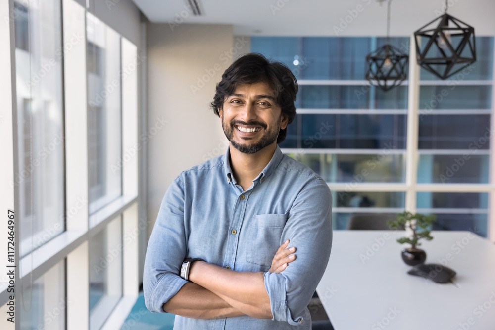 Happy confident Indian company director man standing in office space with arms folded confidence gesture, looking at camera, smiling. Positive businessman, CEO, business owner head shot portrait