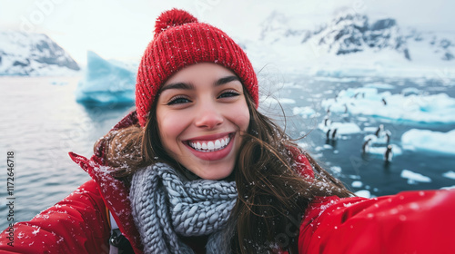 young female traveler in a vibrant red beanie and sport travel jacket, standing on the deck of a ship. She smiles brightly as she takes a selfie, with a group of adorable penguins visible on ice floe