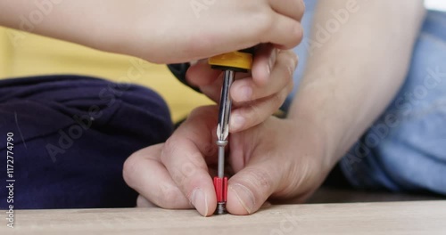 Man and preschooler work to drive screw into wooden surface. Young hands hold screwdriver turning clockwise. Man holds bolt securing detail closeup
