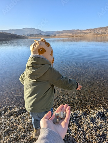A two-year-old boy gleefully throws stones into a clear lake, watching the water ripple with each toss. This playful moment captures the pure joy of childhood exploration and the natural instinct 