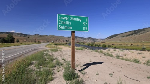 A road sign on Idaho State Highway 75 at Stanley’s entrance shows distances to towns like Challis and Salmon. The Salmon River flows by, cattle graze, and a truck with a trailer passes.