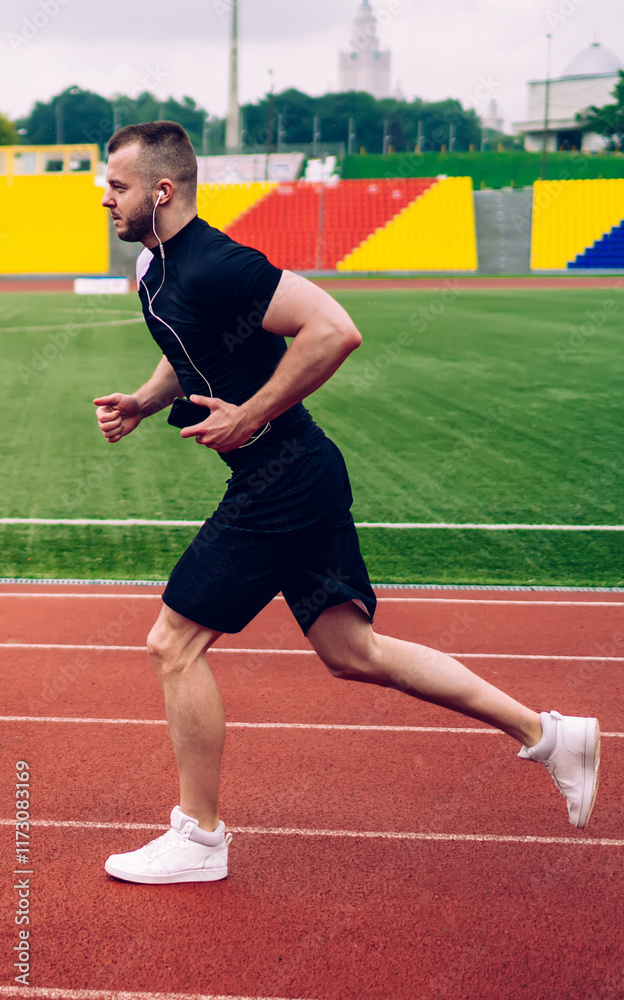 Fototapeta premium Young handsome male in earphones running on stadium dressed in active wear reaching sport goals, strong man athlete keeping fit having morning cardio training with favorite music in headphones