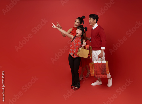 Happy Asian family holding shopping bags and pointing finger isolated on red copy space background. shopping in chinese new year 2025 concept.