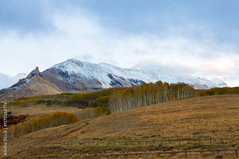 Fototapeta premium Landscape with fenced land and forests of yellowing mixed trees at the foot of snow peaked mountains during hazy fall morning in Colorado, USA