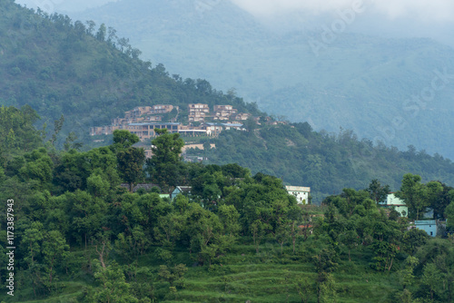A hillside view of a cluster of modern buildings on a slope, surrounded by lush green vegetation and smaller houses below. The misty mountains in the background add a serene atmosphere in uttarakhand.