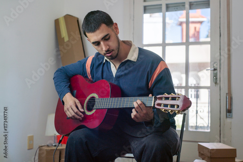 Young man playing red acoustic guitar sitting on chair in his new apartment