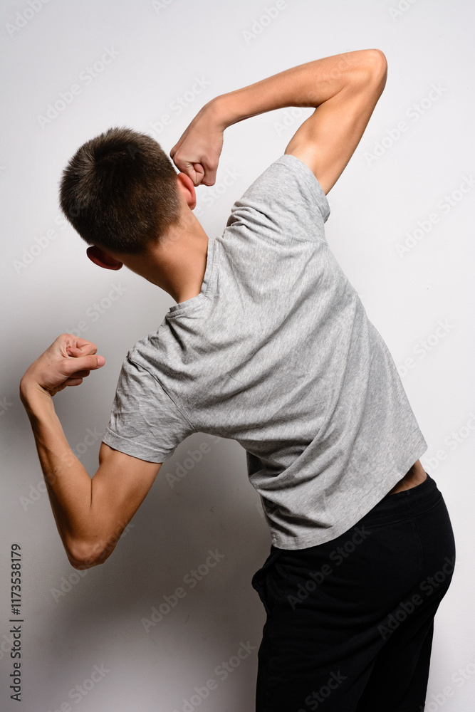 A rear view of a young man in a grey t-shirt and black pants flexing his biceps. He's posed against a plain white backdrop.