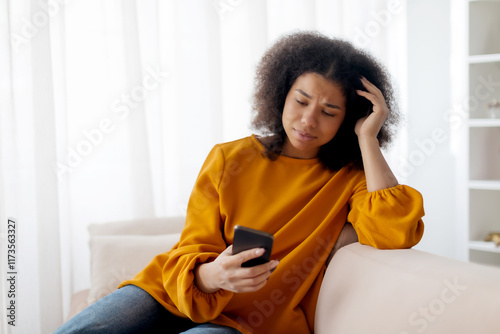 Worried african lady looking at smartphone screen, sitting on couch at home, concerned african american woman waiting for important call, reading unpleasant sms, spending time with phone in living
