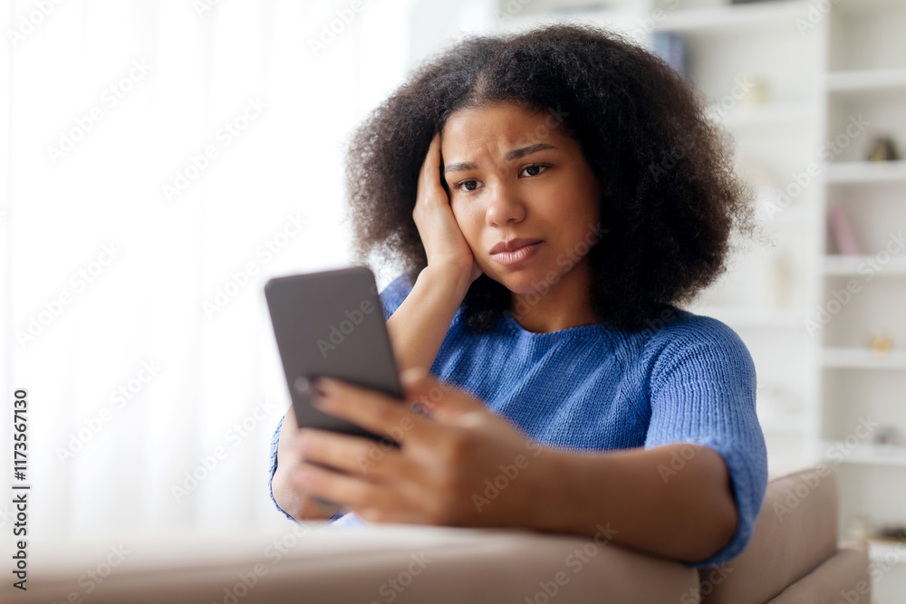 Concerned young african american woman looking at smartphone screen and touching head, upset african american female sitting on couch with troubled expression, suffering stress or got bad news on Concerned young african american woman looking at smartphone screen and touching head, upset african american female sitting on couch with troubled expression, suffering stress or got bad news on