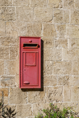 An old red wooden mailbox mounted on a stone wall, with faded paint and weathered edges. Its rustic charm and vintage character stand out against the textured stone backdrop, evoking a sense of nostal photo