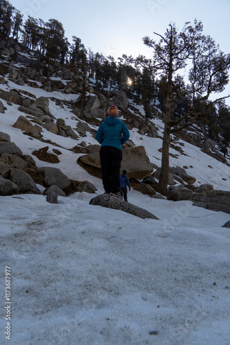 A woman in blue jacket and colorful hat stands on a snowy slope facing a rocky mountain with tall pine trees. Another person walks ahead near a lone tree. The sun peeks through the forest in himachal. photo