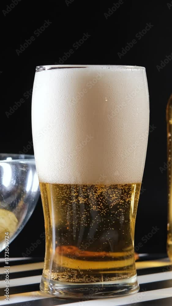 Vertical video. Chilled beer with frothy foam and bubbles near crispy snacks on dark backdrop