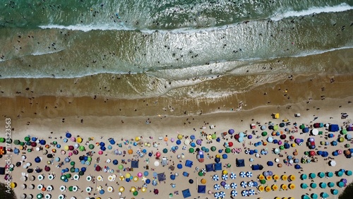 Overhead aerial drone photo of Peró Beach, Cabo Frio, Rio de Janeiro, Brazil. The view highlights colorful umbrellas, golden sand, and ocean waves in a vibrant summer scene.