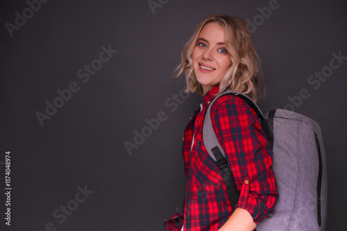 A smiling young woman in red plaid shirt with a gray backpack poses against a dark gray background, exuding confidence and readiness for travel or adventure. Perfect for lifestyle or travel concepts photo