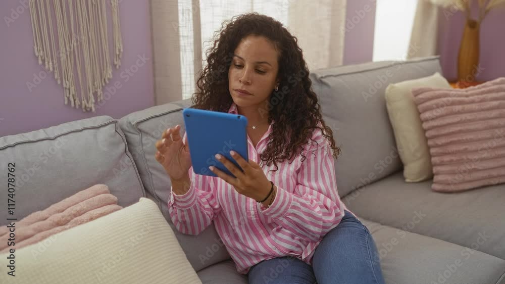 Young woman with curly hair using tablet in cozy living room, wearing pink striped shirt, relaxing on a modern sofa with pastel pillows and macrame wall hanging Young woman with curly hair using tablet in cozy living room, wearing pink striped shirt, relaxing on a modern sofa with pastel pillows and macrame wall hanging