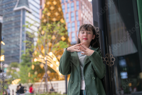 A young girl in a green coat posing gracefully in a festive urban setting, with a glowing Christmas tree and sparkling lights in the background, capturing a serene and joyful holiday moment. photo