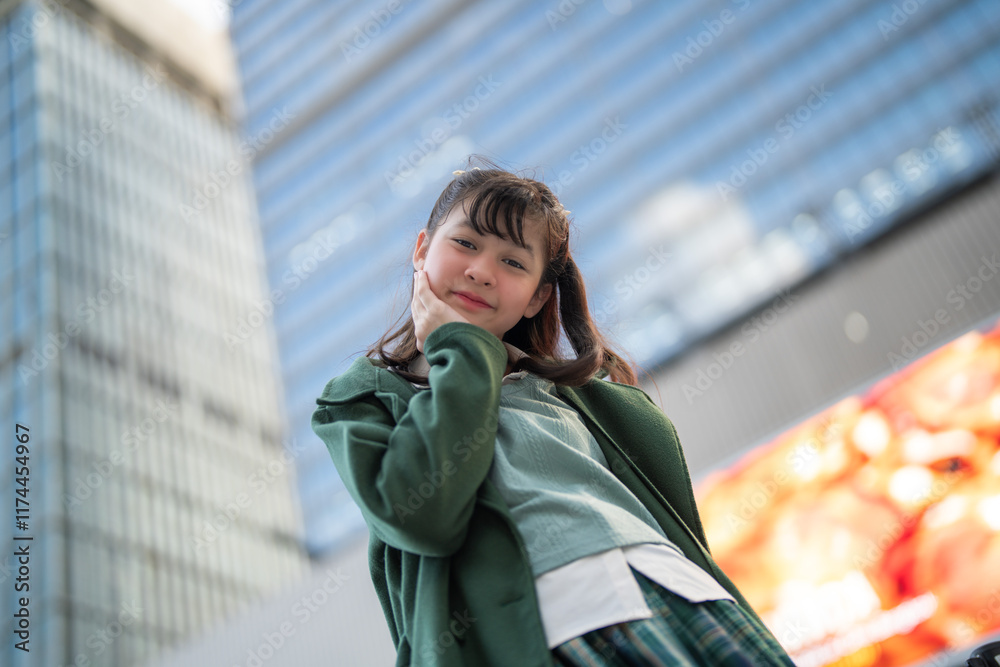 Fototapeta premium A confident young woman in a green coat standing outdoors, looking upward against the backdrop of a modern glass building, symbolizing ambition, contemporary style, and urban living.