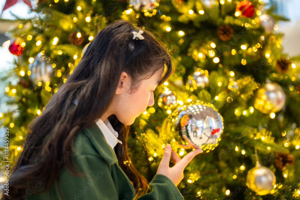 Naklejka premium A young girl in a green coat admiring a reflective ornament on a beautifully decorated Christmas tree, surrounded by glowing lights and festive holiday decorations, capturing a magical moment.