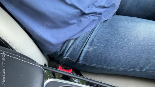 A person is sitting in a car with their leg up on the dashboard and fasten the seatbelt before driving. The person is wearing a blue shirt and blue jeans