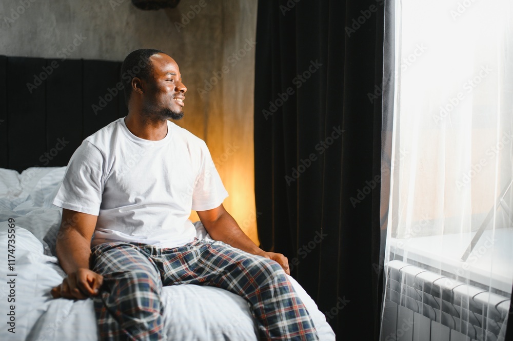 Fototapeta premium African american man wearing pajamas sitting on bed after waking up in the morning. Comfortable healthy sleep concept