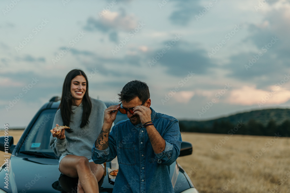 Fototapeta premium Romantic couple leaning on a car, sharing pizza while watching a golden sunset in a meadow