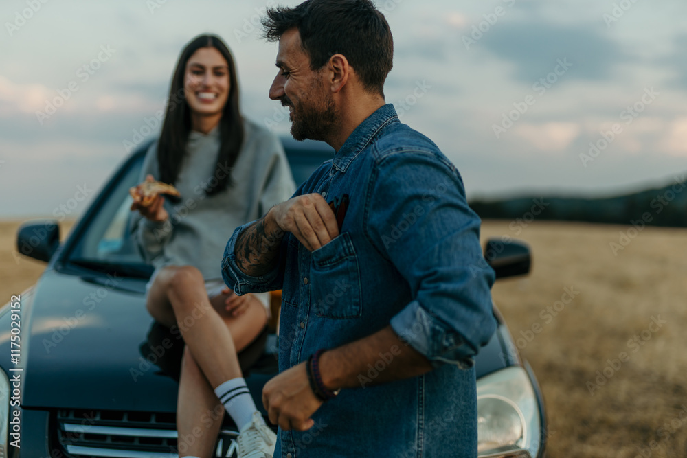 Romantic couple leaning on a car, sharing pizza while watching a golden sunset in a meadow Romantic couple leaning on a car, sharing pizza while watching a golden sunset in a meadow