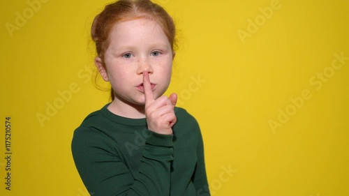 Young girl with red hair gestures for silence against a bright yellow backdrop in a silent moment
