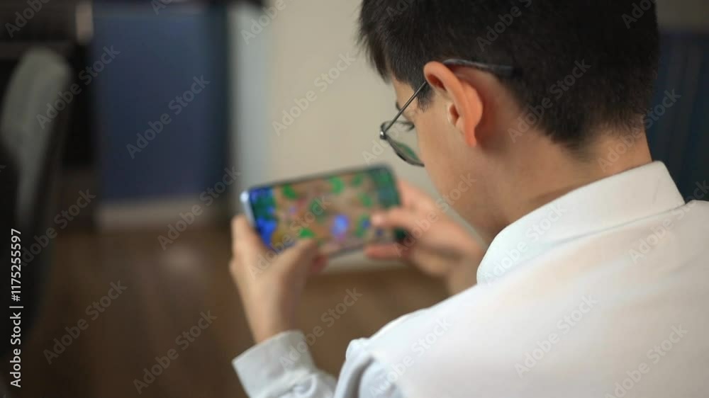 Close up of unrecognized young boy engaged in mobile game while sitting at home. Teenager focuses on screen using smartphone. Back view of kid in glasses enjoying online game through mobile app. Close up of unrecognized young boy engaged in mobile game while sitting at home. Teenager focuses on screen using smartphone. Back view of kid in glasses enjoying online game through mobile app.
