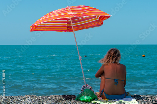 A girl in a blue swimsuit sits in the shade of a red umbrella on the seashore. photo