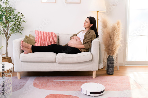 Pregnant woman relaxing in living room while Robotic vacuum cleaner working.