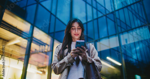 Cute Caucasian girl finishing entering text on her smartphone. Lowering mobile device while glancing at camera. Smiling with joy. Tall building with glass windows in background.