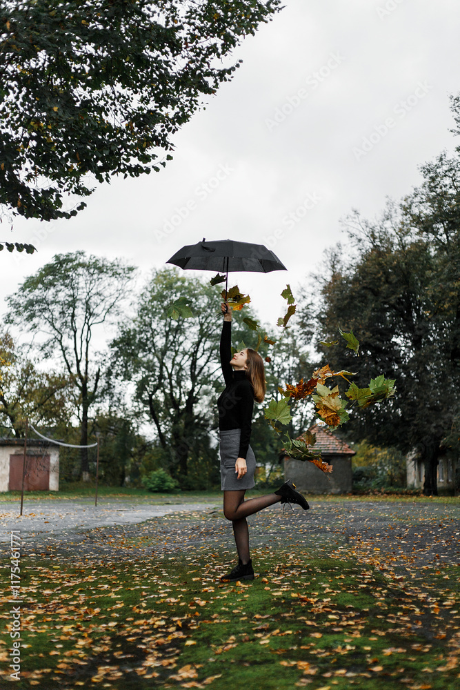 girl standing on one leg on the street in an autumn park. She is wearing a black sweater, a gray skirt and black tights. In one hand she holds a black umbrella raised up. girl standing on one leg on the street in an autumn park. She is wearing a black sweater, a gray skirt and black tights. In one hand she holds a black umbrella raised up.