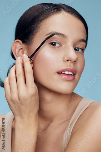 A young woman applying eyebrow gel, showcasing a natural beauty look with a soft smile against a blue background, perfect for cosmetics and skincare promotions