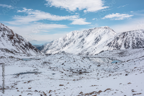 Cold snow-white landscape of snowy valley with alpine lakes against snow-covered rocky mountain range under clouds in blue sky. Glacial lakes, pure white snow and stones in high mountains in sunny day