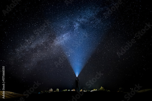 Stunning night panorama in the Moroccan desert. A man illuminates the sky with his flashlight, merging the light with the Milky Way. In the background there is a small illuminated camp