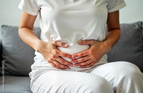 Young Asian woman sits on couch holding stomach. Appears to in pain. Possible causes include food poisoning, digestive issues, menstrual cramps. Experiencing abdominal discomfort. Hands on belly.