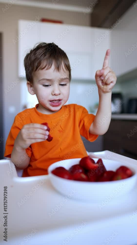 Adorable baby boy in orange t-shirt sits in front of the big bowl of strawberries. Kid eats and counts red berries. Close up. Vertical video. Adorable baby boy in orange t-shirt sits in front of the big bowl of strawberries. Kid eats and counts red berries. Close up. Vertical video.
