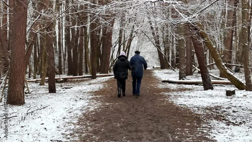 Mature couple going on snowy path at winter pine forest. Senior husband and wife walking through trail at coniferous woodland. Older pair of pensioners spending time together at nature. Slow motion